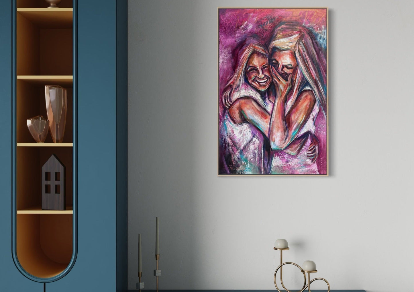 A colorful framed painting of two embracing women hangs on a light gray wall in a modern interior setting with a blue cabinet and decorative objects.