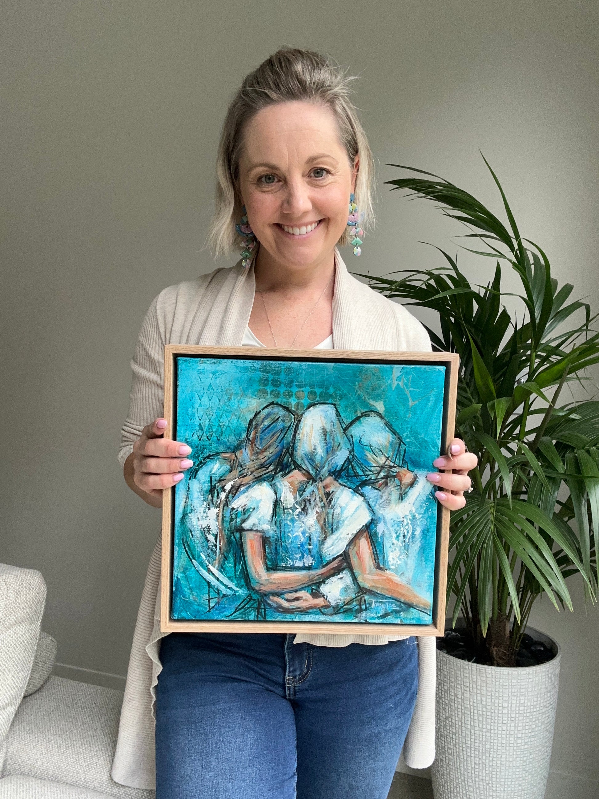 Tracy Lee holding a framed painting of three women embracing, “I stand with you” showing a smile as she presents the artwork in an indoor setting with a potted plant in the background.
