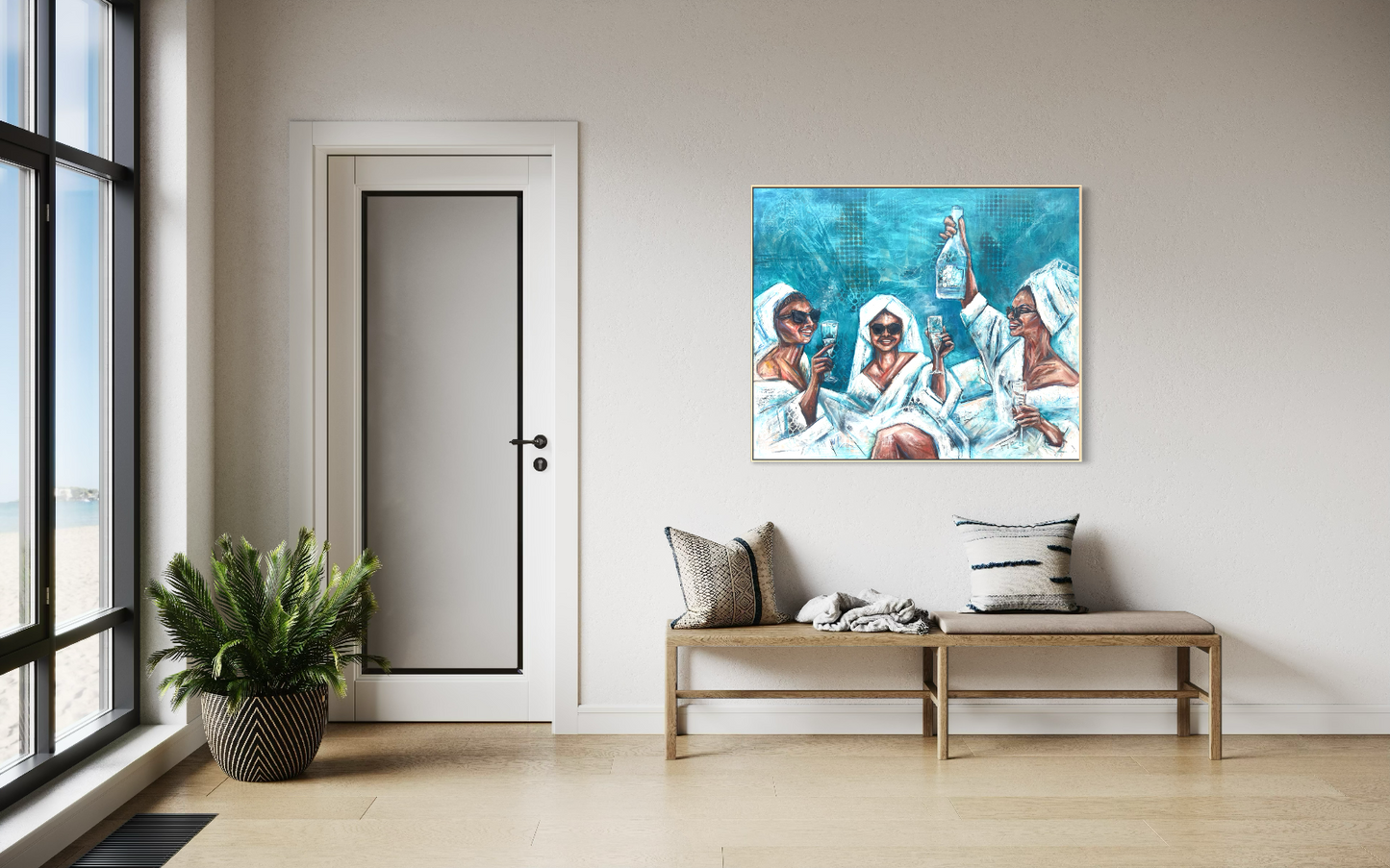 A framed art print depicting four Black women in white robes holding wine glasses, displayed on a light beige wall in a modern, sunlit room with a wooden bench and a potted plant.