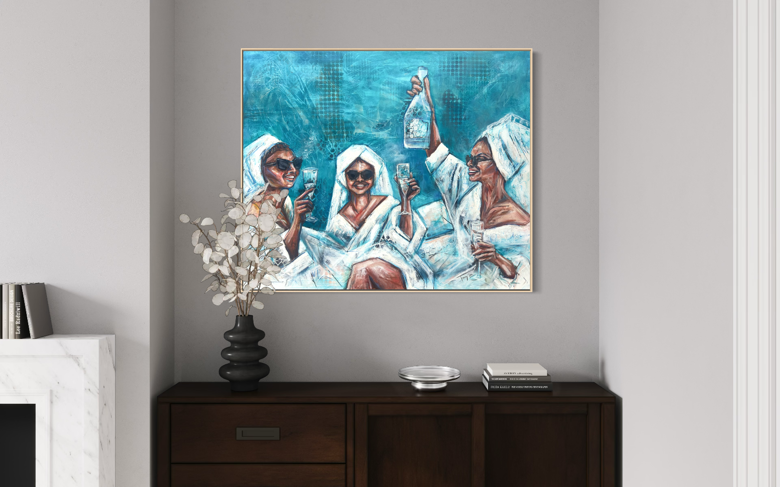 A framed canvas art depicting three women in white robes and sunglasses holding wine glasses, hung on a light grey wall above a dark wooden console table with a vase and books.