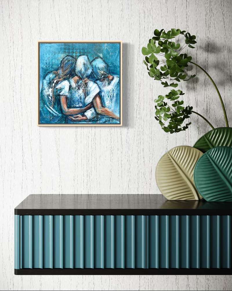 A framed square painting depicting three figures embracing in shades of blue and white, hanging on a white textured wall above a dark console table with decorative leaf-shaped vases and green trailing plants.