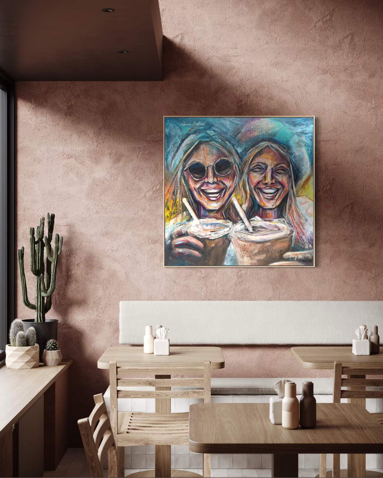 A colorful framed painting of two smiling women holding coffee cups hangs on a textured dusty pink wall in a modern cafe setting with wooden tables and chairs.
