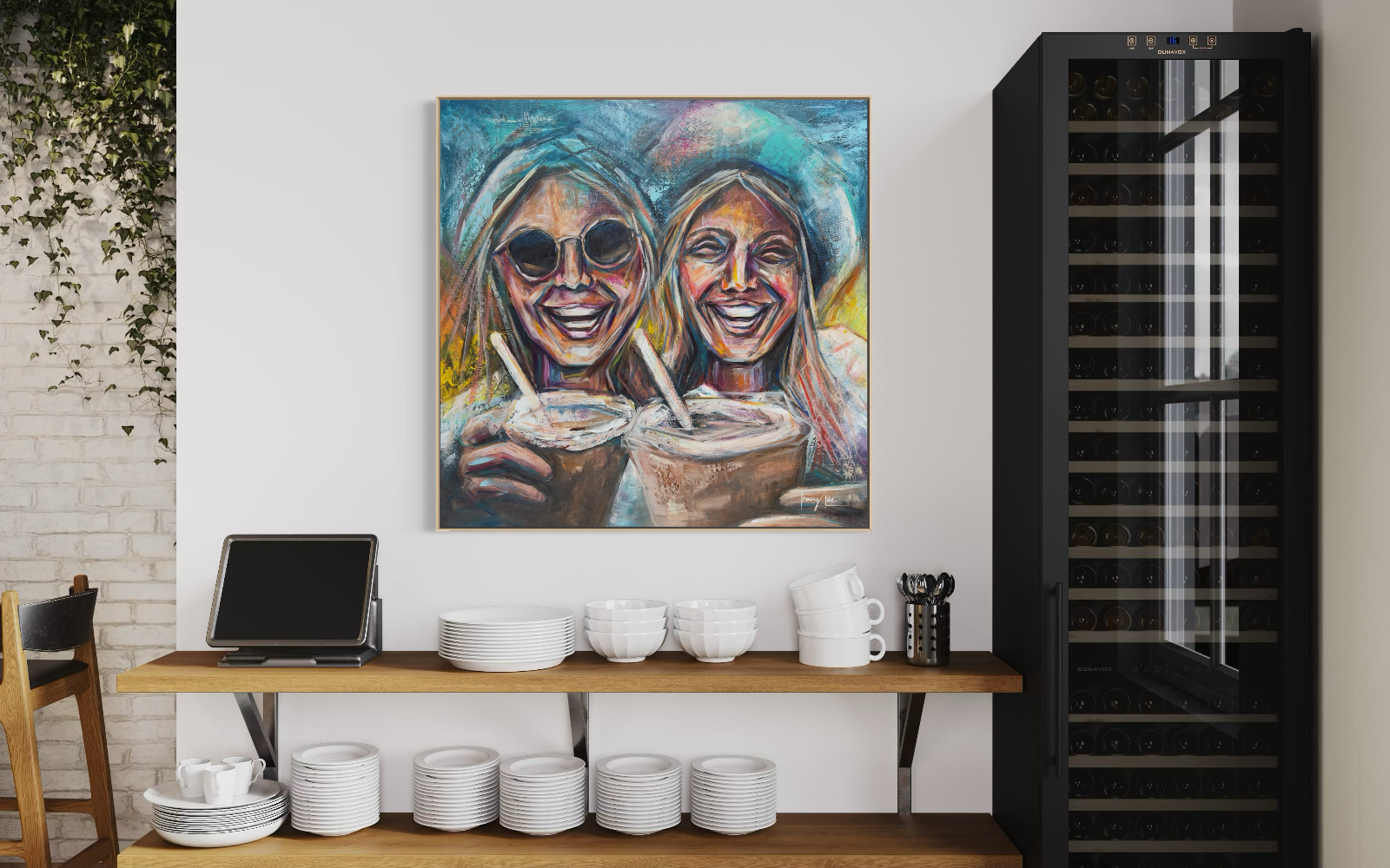 A colorful square painting of two smiling women holding coffee cups hangs on a white wall in a kitchen or dining area. Below the artwork are wooden shelves holding white dishes, mugs, and a small black device, with a dark wine or beverage cabinet to the right.