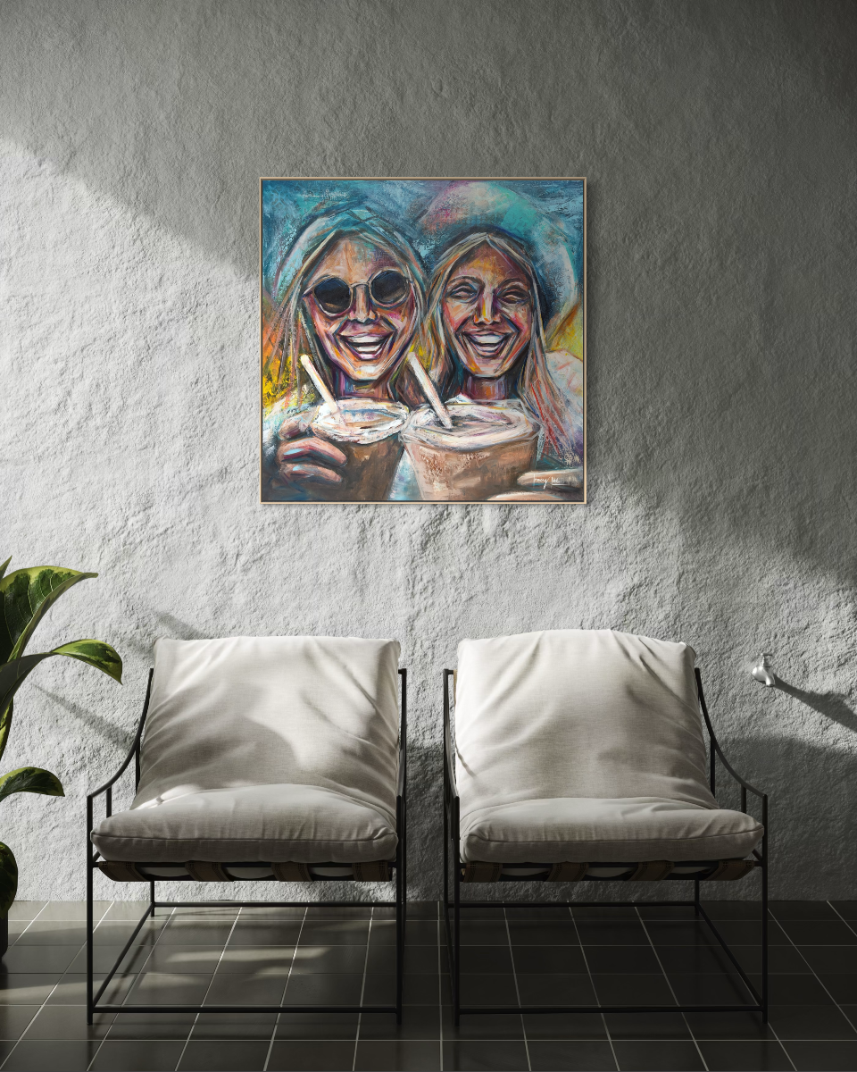 A colorful framed painting of two smiling women holding drinks, displayed on a textured gray wall above two modern armchairs with white cushions in a styled interior setting.