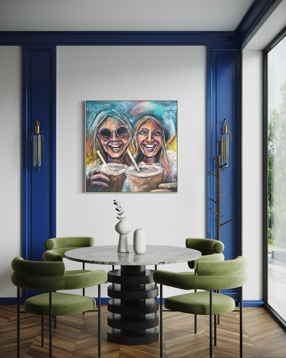 A colorful canvas painting of two smiling women holding drinks, displayed on a white wall in a modern dining room with blue trim, a round gray table, and green chairs.